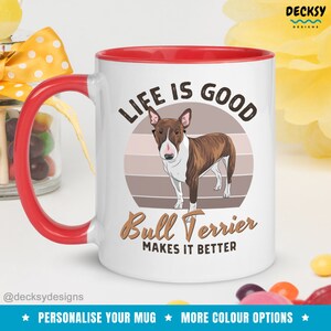 May include: White ceramic mug with a red rim and handle. The mug features a brown and white bull terrier dog illustration with a retro sunset background. The text "Life is good Bull Terrier makes it better" is printed on the mug.