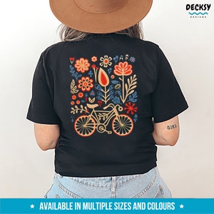 Op de afbeelding: Zwart T-shirt met een kleurrijk bloemenontwerp en een fiets in het midden. Het ontwerp bevat rode, oranje, gele, blauwe en groene bloemen. De tekst "Girl" is gedrukt op de rechtermouw.