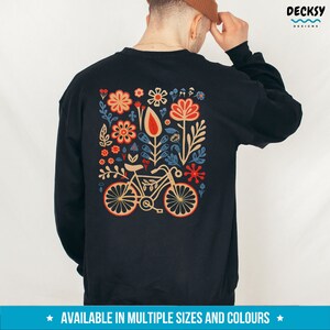 Op de afbeelding: Zwart sweatshirt met een kleurrijk bloemenontwerp en een fiets in het midden. Het ontwerp bevat rode, oranje, gele, blauwe en groene bloemen en bladeren. De tekst "AVAILABLE IN MULTIPLE SIZES AND COLORS" staat onderaan de afbeelding.