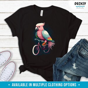 May include: A black t-shirt with a pink and blue cockatoo riding a bicycle. The cockatoo has a pink head and a blue body with yellow accents. The bicycle is red with black tires.