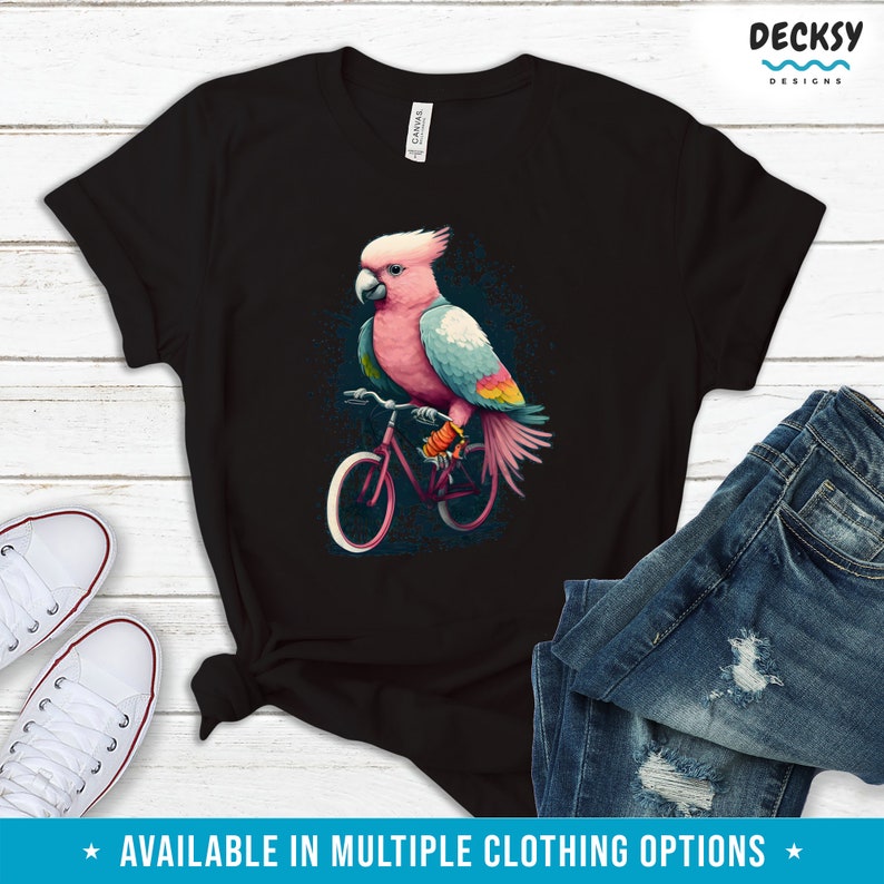 May include: A black t-shirt with a pink and blue cockatoo riding a bicycle. The cockatoo has a pink head and a blue body with yellow accents. The bicycle is red with black tires.