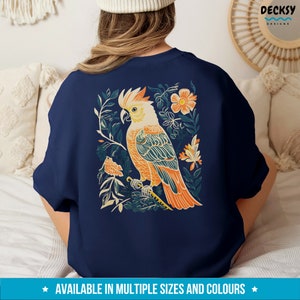 May include: A navy blue sweatshirt featuring a colourful illustration of a cockatoo bird perched on a branch with flowers. The bird is orange, yellow, and blue with a white crest. The flowers are pink, orange, and yellow.