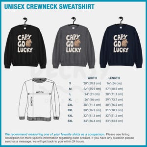 May include: A unisex crewneck sweatshirt with a black, gray, and navy blue color options. The sweatshirt features a graphic of a capybara wearing a cowboy hat with the text "Capy Go Lucky". The size chart shows the width and length measurements in inches and centimeters.