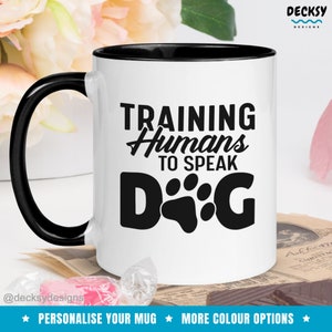 Könnte beinhalten: Weiße Keramik-Kaffeetasse mit schwarzem Rand und Henkel. Die Tasse hat ein schwarzes Motiv mit der Aufschrift "Training Humans to Speak Dog" mit einem Pfotenabdruck.