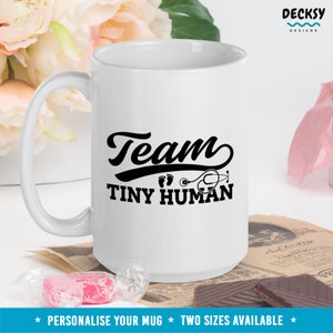 K&ouml;nnte beinhalten: Eine wei&szlig;e Keramiktasse mit einem schwarzen Design, auf dem "Team Tiny Human" mit einem Stethoskop und Fu&szlig;abdr&uuml;cken steht. Die Tasse steht auf einem Tisch mit Schokolade und S&uuml;&szlig;igkeiten.