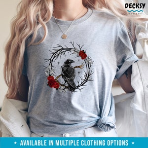 May include: A light gray t-shirt featuring a black crow perched on a branch within a circular wreath of branches, red roses, and white leaves. The shirt has the text "AVAILABLE IN MULTIPLE CLOTHING OPTIONS" at the bottom.