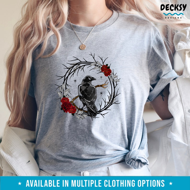 May include: A light gray t-shirt featuring a black crow perched on a branch within a circular wreath of branches, red roses, and white leaves. The shirt has the text "AVAILABLE IN MULTIPLE CLOTHING OPTIONS" at the bottom.