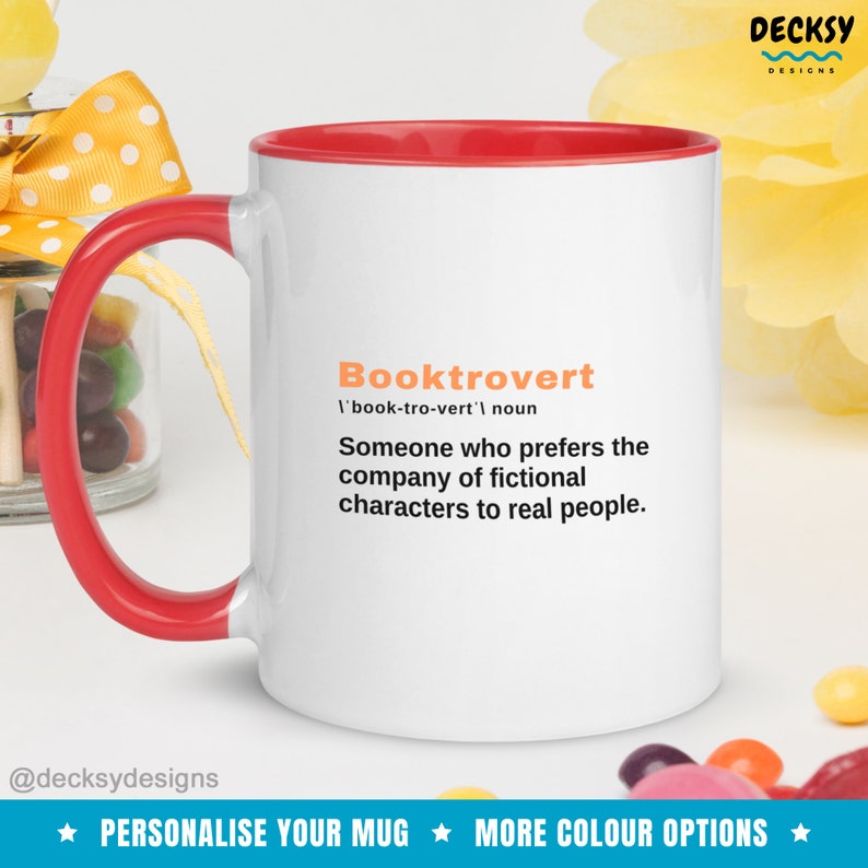 May include: White ceramic mug with a red rim. The mug features a definition of the word "Booktrovert" in black text with an orange accent. The text reads: "Booktrovert /'book-tro-vert'/ noun Someone who prefers the company of fictional characters to real people."