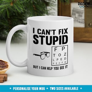 Op de afbeelding: Witte keramische mok met de zwarte tekst "I CAN'T FIX STUPID", een oogtestkaart en een bril. De mok bevat ook de zin "BUT I CAN HELP YOU SEE IT".