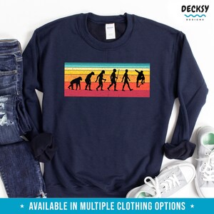 May include: Navy blue sweatshirt featuring a retro-style graphic of human evolution on a skateboard, set against a striped background of orange, yellow, and red. The text "DECKSY DESIGNS" is in the upper right corner. The bottom of the image has the text "AVAILABLE IN MULTIPLE CLOTHING OPTIONS".