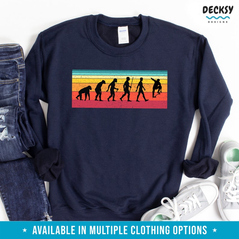 May include: Navy blue sweatshirt featuring a retro-style graphic of human evolution on a skateboard, set against a striped background of orange, yellow, and red. The text "DECKSY DESIGNS" is in the upper right corner. The bottom of the image has the text "AVAILABLE IN MULTIPLE CLOTHING OPTIONS".