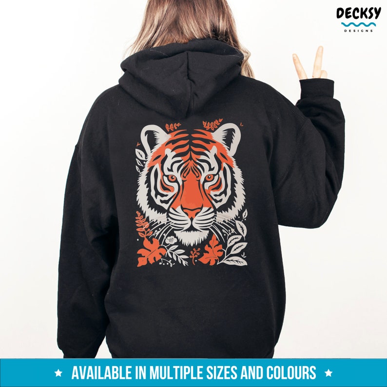 Puede incluir: Sudadera con capucha negra con un gr&aacute;fico de cara de tigre blanco y naranja en la parte posterior. El tigre est&aacute; rodeado de elementos florales. El texto "AVAILABLE IN MULTIPLE SIZES AND COLOURS" est&aacute; en la parte inferior de la imagen.