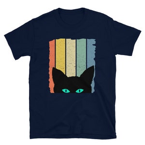 May include: A navy blue t-shirt with a retro-style graphic of a black cat peeking out from behind colorful stripes. The cat has bright green eyes.