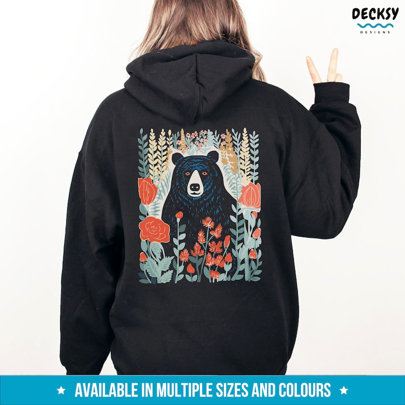 May include: Black hoodie with a graphic print of a black bear surrounded by colorful flowers. The bear is looking directly at the viewer. The text "AVAILABLE IN MULTIPLE SIZES AND COLORS" is printed at the bottom of the image.