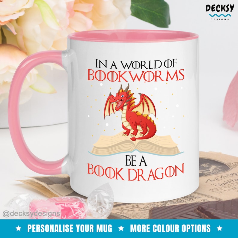 May include: White ceramic mug with a pink rim and handle. The mug features a red dragon sitting on an open book with the text "In a world of bookworms be a book dragon".