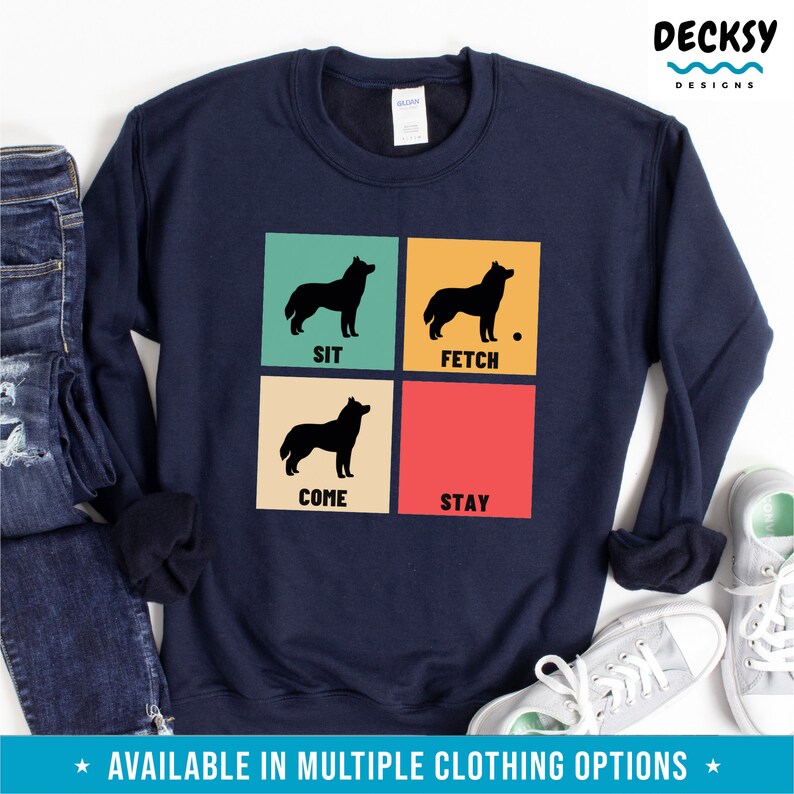 Op de afbeelding: Een marineblauw sweatshirt met een retro-ontwerp met vier zwarte silhouetten van een husky-hond met de woorden "Sit", "Fetch", "Come" en "Stay" in witte tekst.