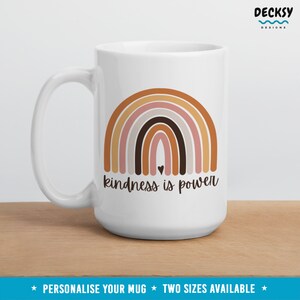 May include: White ceramic mug with a rainbow design in shades of brown, pink, and gray. The text "kindness is power" is printed below the rainbow. A small heart is in the center of the rainbow.