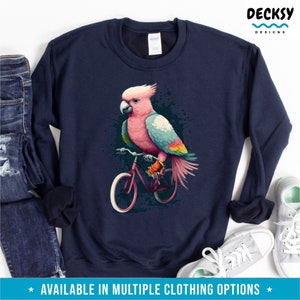 May include: A navy blue sweatshirt with a pink and blue cockatoo riding a red bicycle. The cockatoo has a white crest and a yellow beak. The bicycle has white tires and a black frame. The background is a dark blue with white splatters.
