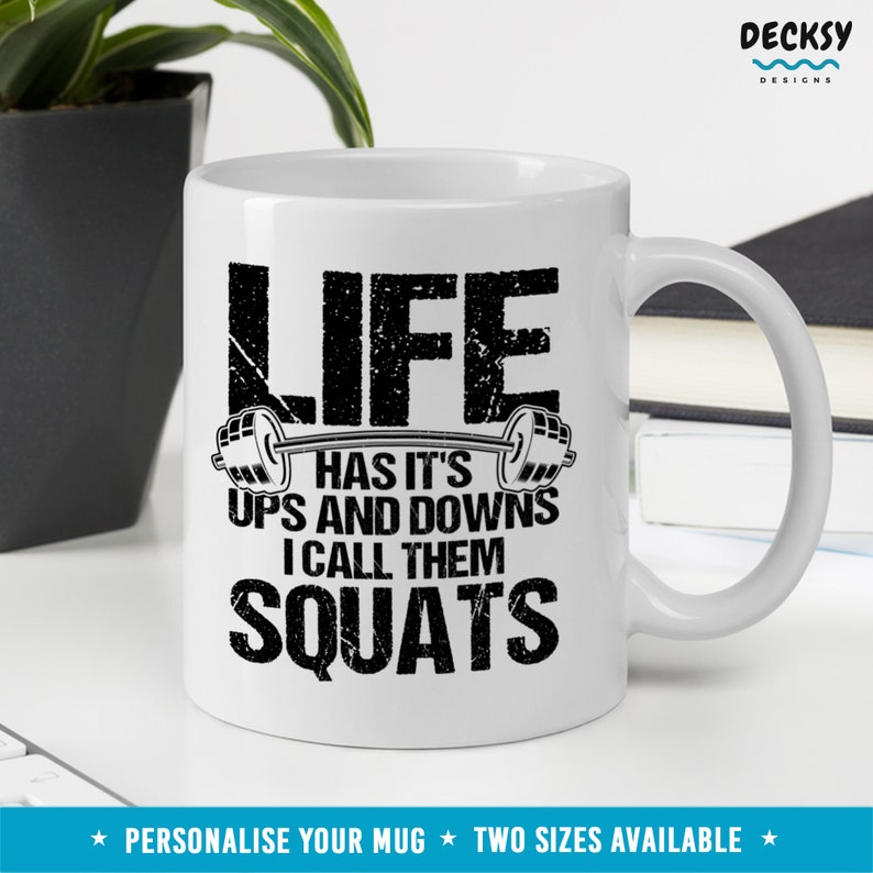 May include: White ceramic mug with a black graphic that reads "Life has it's ups and downs I call them squats" with a barbell and weights on either side of the text.