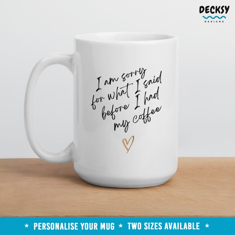 May include: White ceramic coffee mug with black text that reads "I am sorry for what I said before I had my coffee" with a gold heart below the text.  The mug is on a wooden surface.