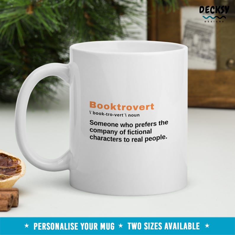 May include: White ceramic mug with a black and orange design. The mug features the word "Booktrovert" defined as "Someone who prefers the company of fictional characters to real people."