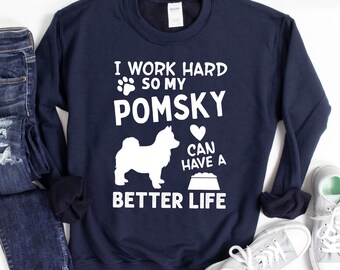 May include: A navy blue sweatshirt with white text that reads "I work hard so my Pomsky can have a better life". The text is accompanied by a white paw print, a white silhouette of a Pomsky dog, a white heart, and a white bowl. The text "AVAILABLE IN MULTIPLE CLOTHING OPTIONS" is at the bottom of the sweatshirt.