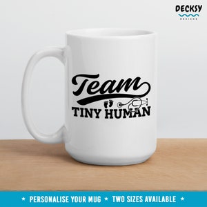 K&ouml;nnte beinhalten: Wei&szlig;e Keramiktasse mit einem schwarzen Design, auf dem "Team Tiny Human" mit einem Stethoskop und Fu&szlig;abdr&uuml;cken steht.