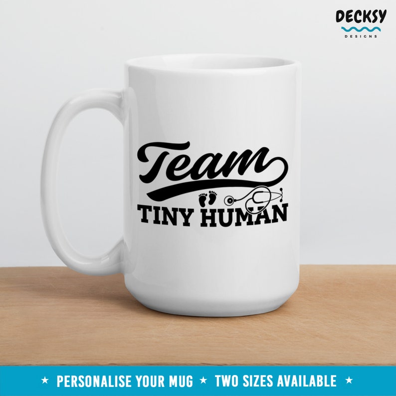 K&ouml;nnte beinhalten: Wei&szlig;e Keramiktasse mit einem schwarzen Design, auf dem "Team Tiny Human" mit einem Stethoskop und Fu&szlig;abdr&uuml;cken steht.