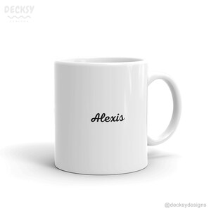 K&ouml;nnte beinhalten: Eine wei&szlig;e Keramiktasse mit einer schwarzen, kursiv geschriebenen Aufschrift "Alexis".