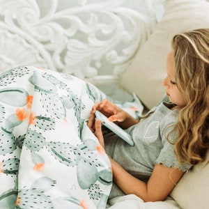 May include: A white blanket featuring a watercolour cactus design in green and orange hues. A person is using a tablet while lying on a bed. The person is wearing a grey t-shirt with a heart motif.