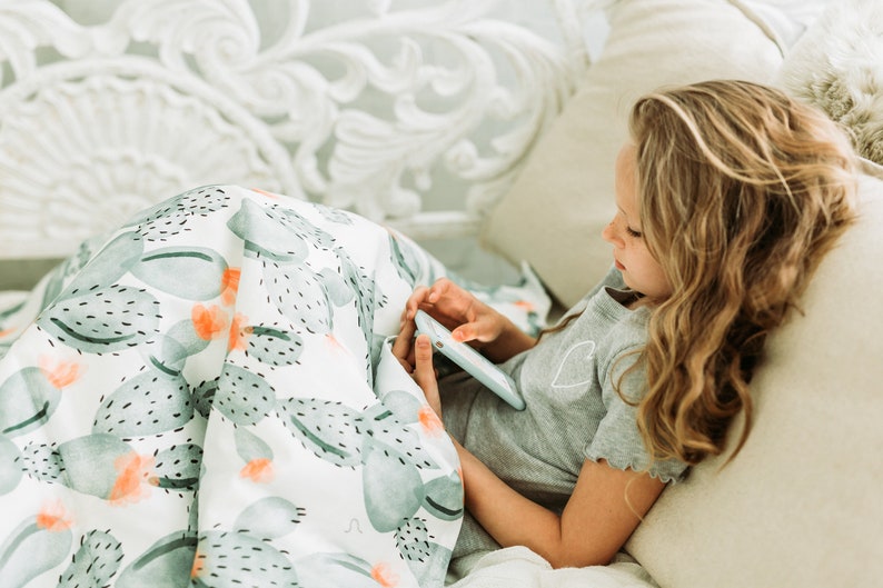 May include: A white blanket featuring a watercolour cactus design in green and orange hues. A person is using a tablet while lying on a bed. The person is wearing a grey t-shirt with a heart motif.