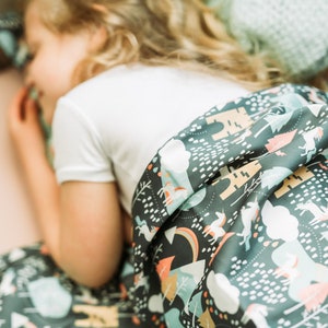 May include: A child asleep under a dark grey blanket with a whimsical forest pattern. The blanket features unicorns, trees, and clouds in white, peach, and teal. The child is wearing a white t-shirt.