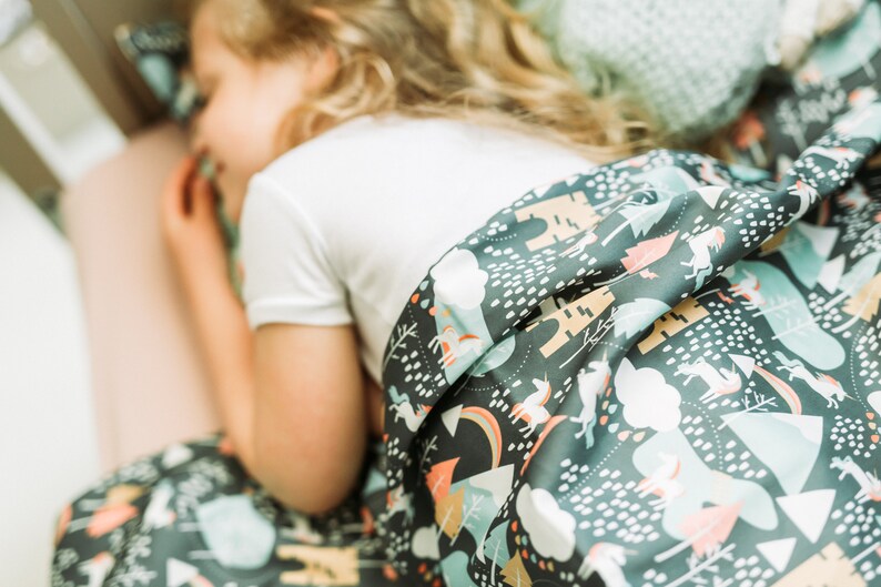 May include: A child asleep under a dark grey blanket with a whimsical forest pattern. The blanket features unicorns, trees, and clouds in white, peach, and teal. The child is wearing a white t-shirt.