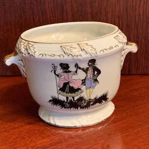 May include: White ceramic planter with gold trim and two handles. The planter features a silhouette of a couple dancing in a garden setting.