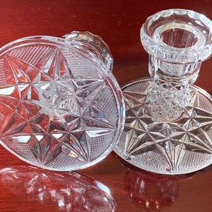 May include: Two clear glass candle holders with a starburst pattern. The holders have a round base and a tall, cylindrical candle cup.
