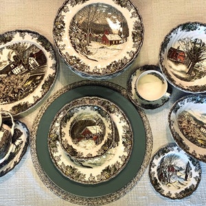 May include: A collection of vintage dinnerware featuring a winter landscape scene in black, white, and red. The set includes plates, bowls, a cup and saucer, and an oval serving dish. The rims are decorated with a floral pattern.