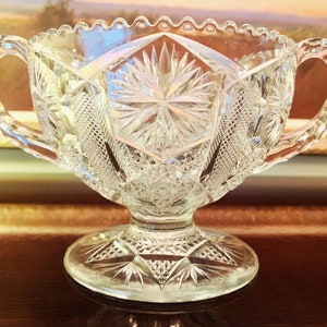 May include: A clear glass sugar bowl with two handles and a pedestal base. The bowl has a cut glass design with a starburst pattern.
