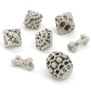 May include: A set of seven bone-shaped dice with skull and crossbones designs. The dice are white with black markings and include a d4, d6, d8, d10, d%, d12, and d20.