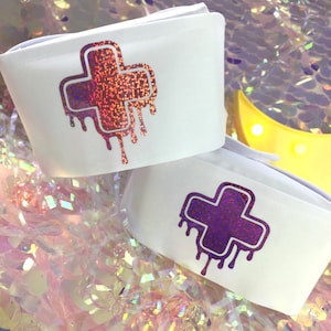 May include: Two white fabric wrist cuffs with a dripping plus sign design. The plus sign on the top cuff is iridescent pink and orange. The plus sign on the bottom cuff is iridescent purple.
