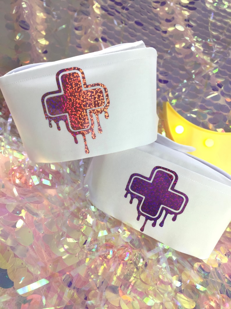 May include: Two white fabric wrist cuffs with a dripping plus sign design. The plus sign on the top cuff is iridescent pink and orange. The plus sign on the bottom cuff is iridescent purple.