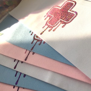 May include: Stack of white and light blue fabric squares with a pink glittery dripping cross design.