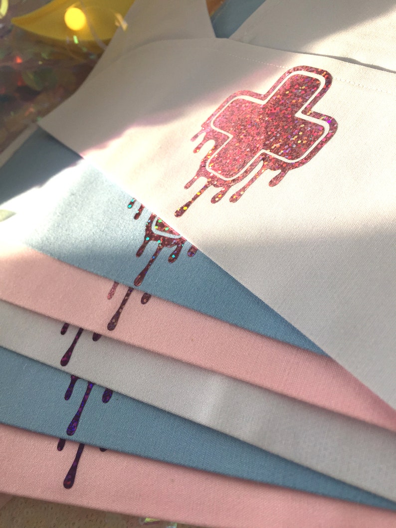 May include: Stack of white and light blue fabric squares with a pink glittery dripping cross design.
