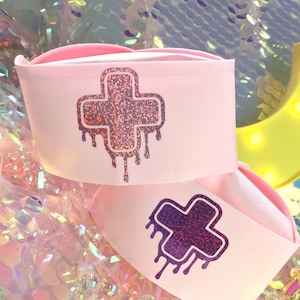 May include: Two pink nurse hats with a glittery dripping cross design. The cross on the top hat is pink and the cross on the bottom hat is purple.