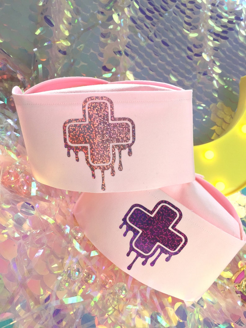 May include: Two pink nurse hats with a glittery dripping cross design. The cross on the top hat is pink and the cross on the bottom hat is purple.