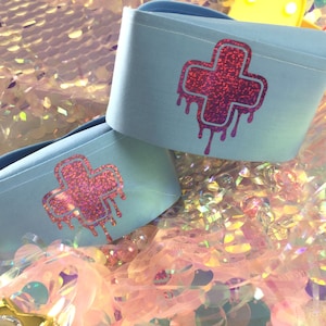 May include: Two light blue fabric wrist cuffs with a pink glitter dripping cross design.