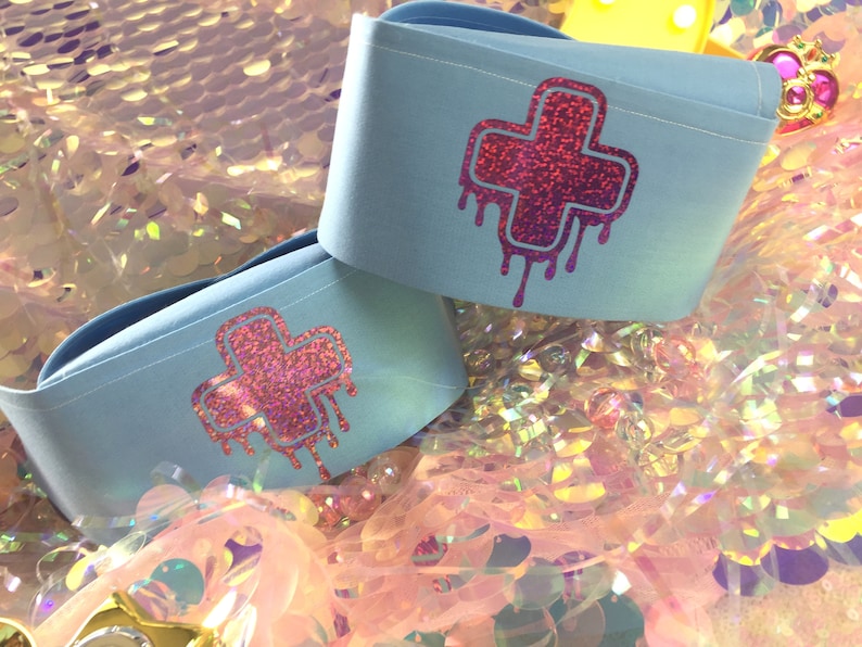 May include: Two light blue fabric wrist cuffs with a pink glitter dripping cross design.