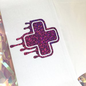 May include: A white fabric patch with a pink and purple glitter plus sign with a white outline. The plus sign has a dripping effect around the edges.