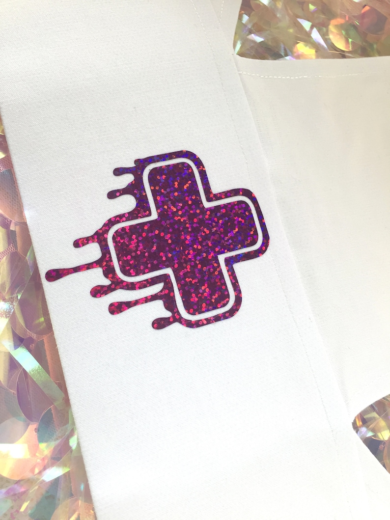 May include: A white fabric patch with a pink and purple glitter plus sign with a white outline. The plus sign has a dripping effect around the edges.