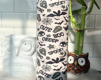May include: A white tumbler with a black and gray Halloween-themed design. The design includes bats, skulls, spiderwebs, crosses, and the words "Cute Boo Creep", "Spooky", and "Ouija".