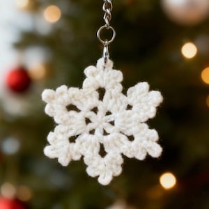 Op de afbeelding: Een witte, gehaakte sneeuwvlok sleutelhanger hangend aan een tak. De sneeuwvlok heeft gedetailleerd openwerk, en de sleutelhanger heeft een zilverkleurige sluiting en ketting. De achtergrond is een vage kerstboom met ornamenten.